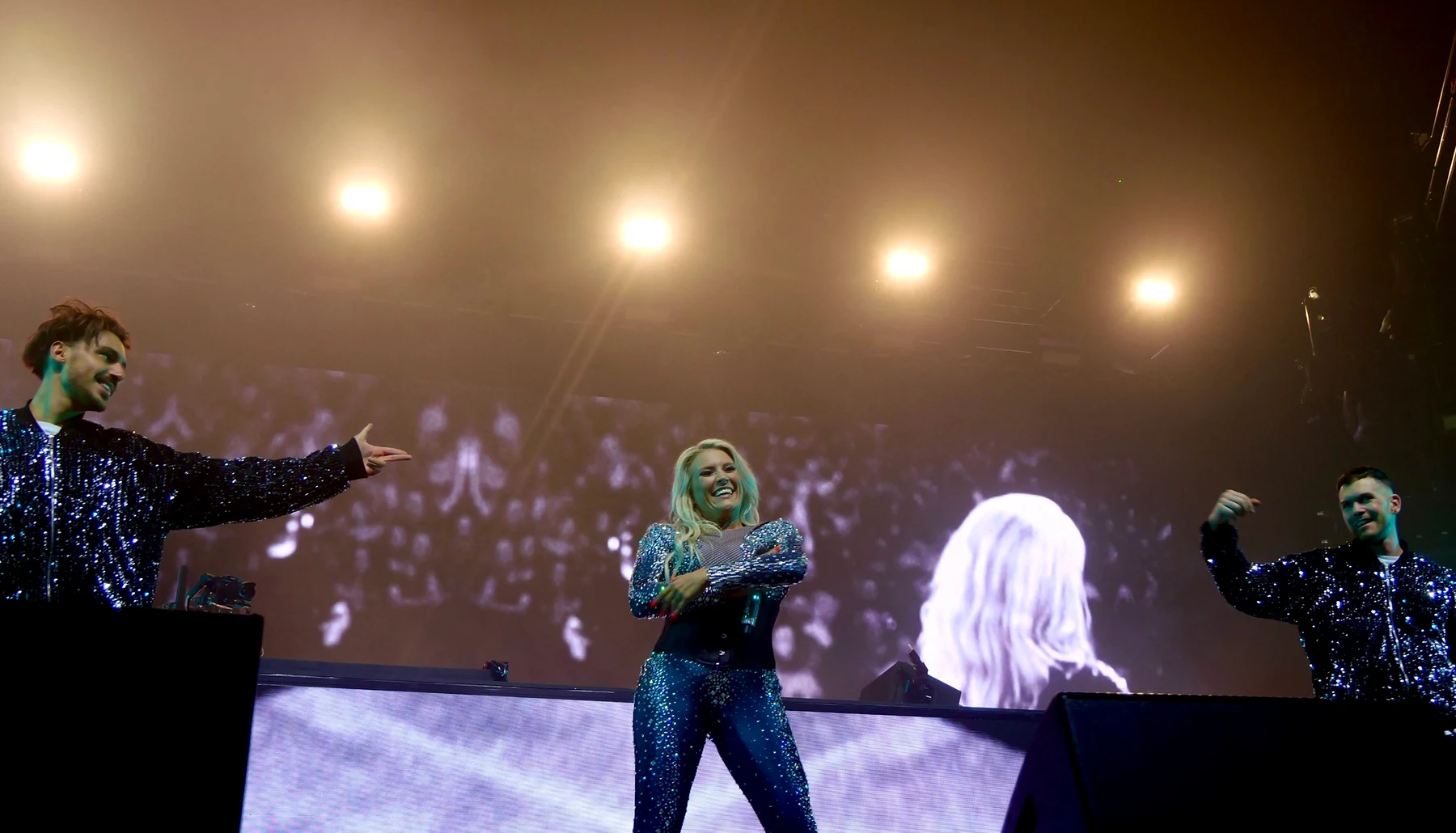 Cascada laughing on stage between two dancers in sequinned jackets, under warm stage lights