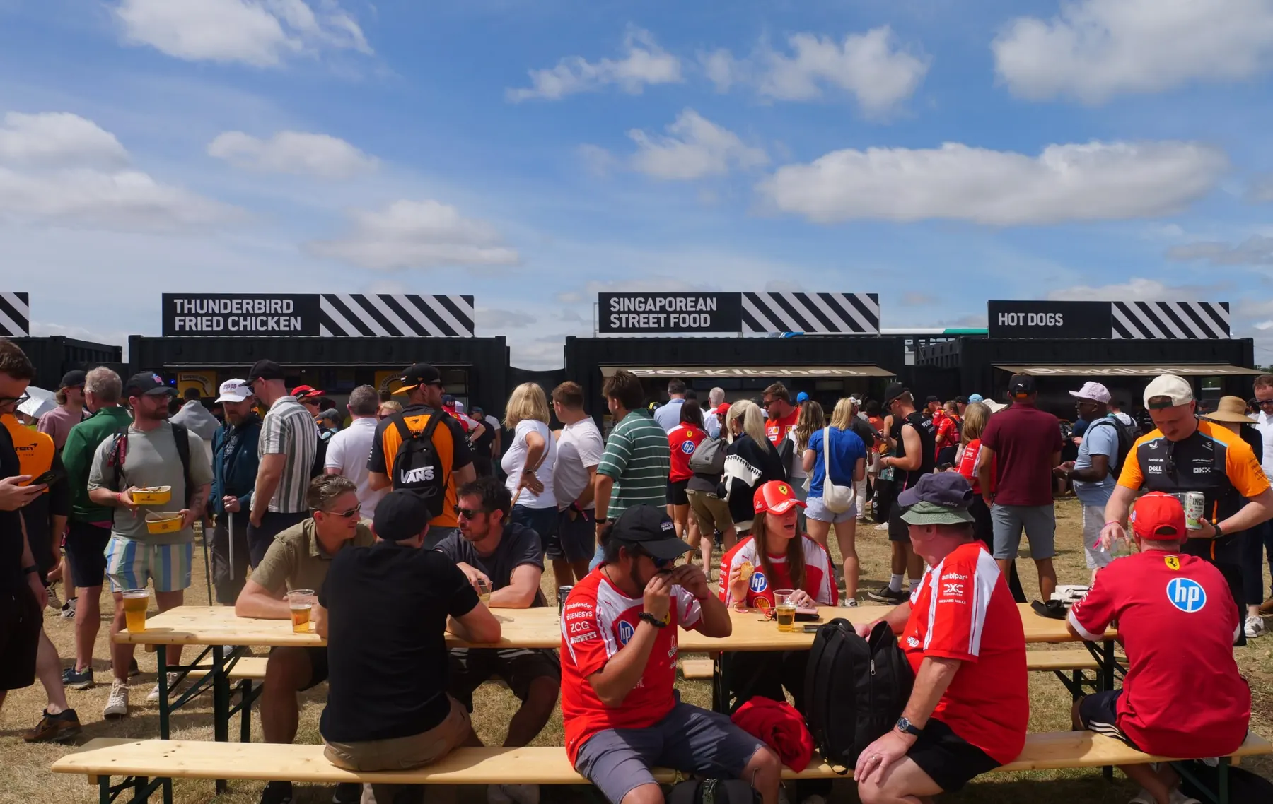 Formula 1 fans in team shirts eating at benches in front of black BoxKitchen container kitchens at Silverstone during the British Grand Prix