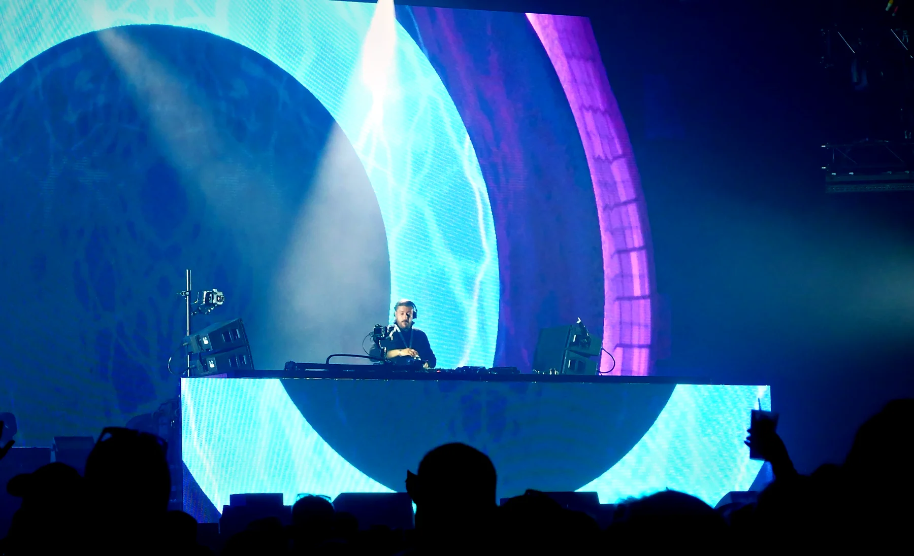 DJ at a glowing booth in front of a huge cyan orb, with white spotlights cutting through the haze and hands up in the crowd