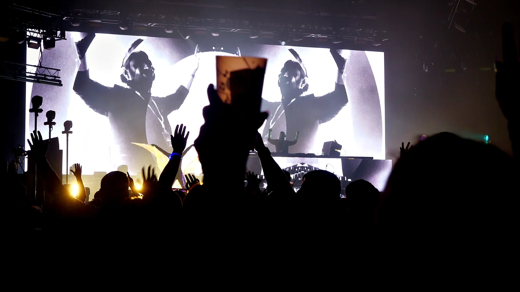Crowd with hands and a pint raised in silhouette in front of a giant black and white screen of the DJ with arms up