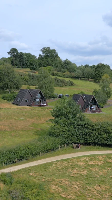 Drone shot of two dark timber A-frame holiday cabins on a green hillside near a lake outside Nottingham, a gravel path and hedgerow in the foreground