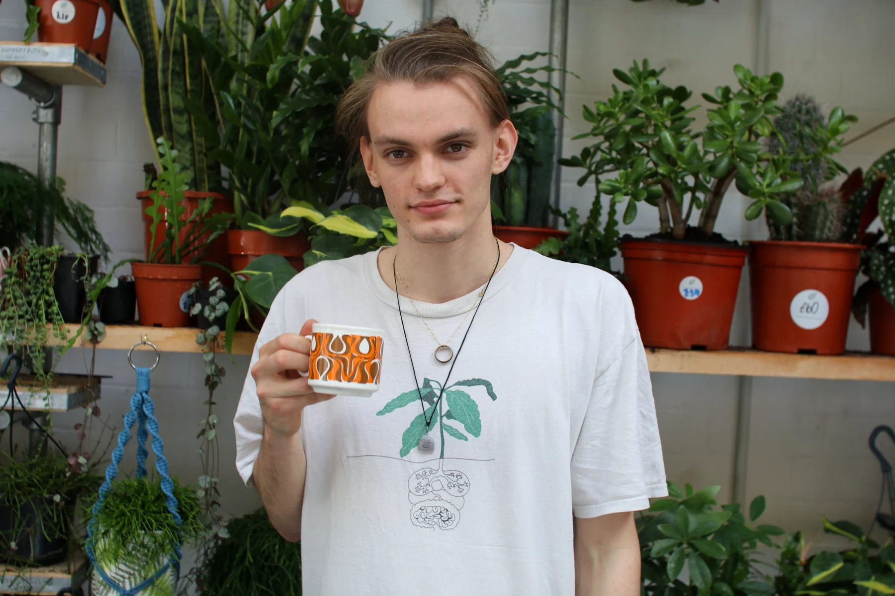 The Lono Espresso barista holding up an orange retro patterned cup in front of shelves of potted plants