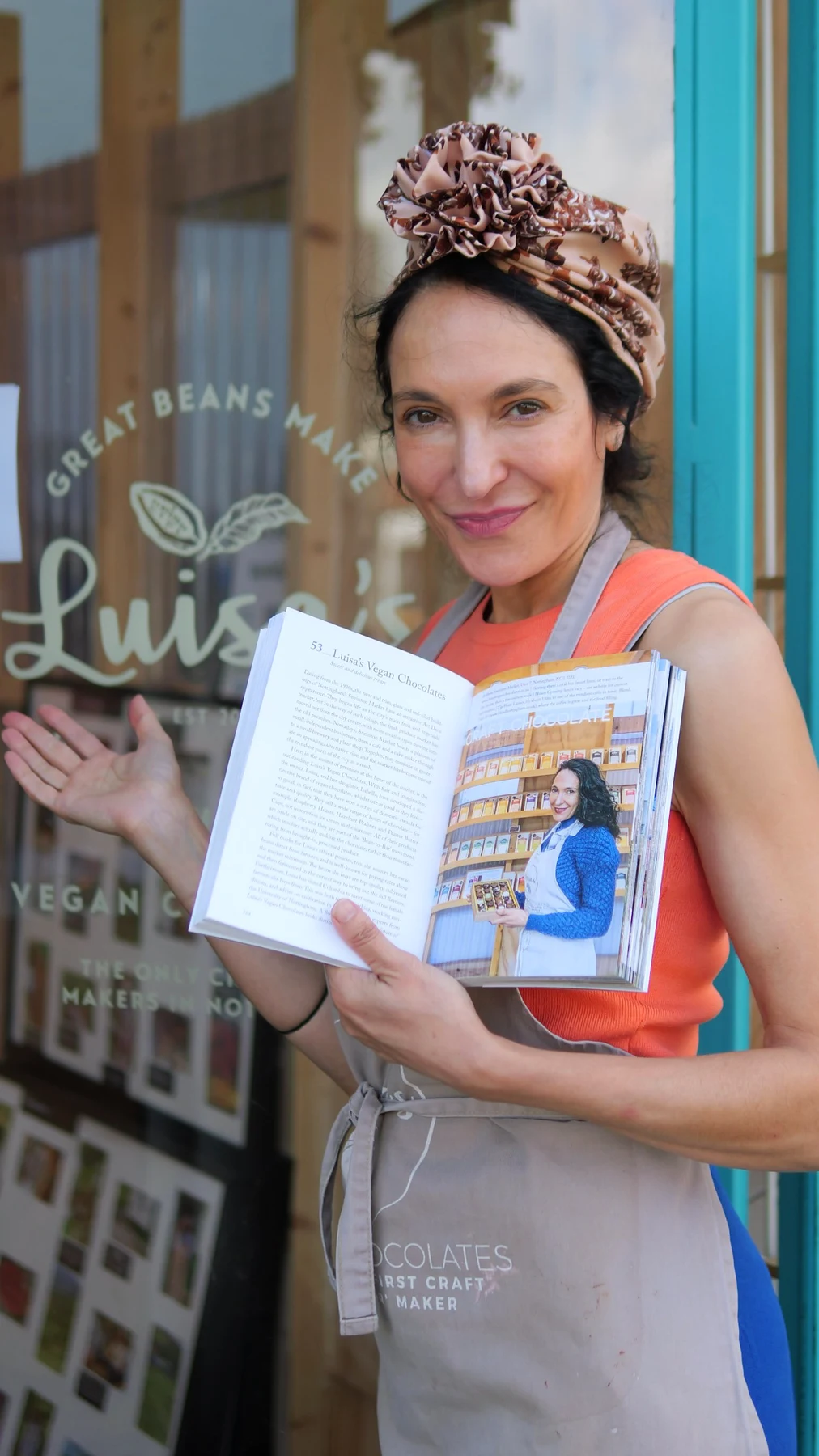 Chocolatier in a Luisa's apron outside the shop window, holding open a Nottingham guide book at the page featuring Luisa's Vegan Chocolates