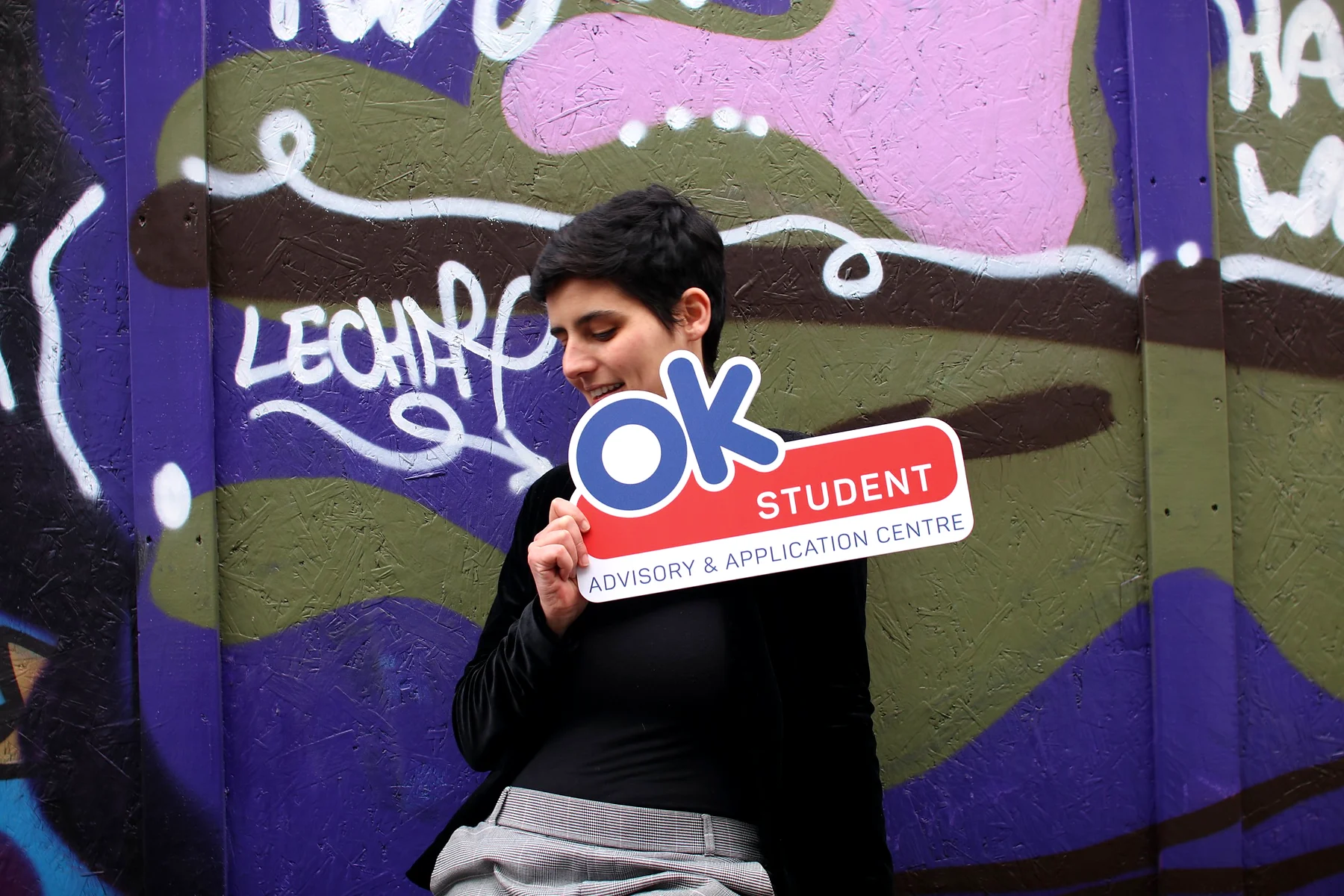 A student with short dark hair smiling down at an OK Student sign in front of a purple graffiti wall