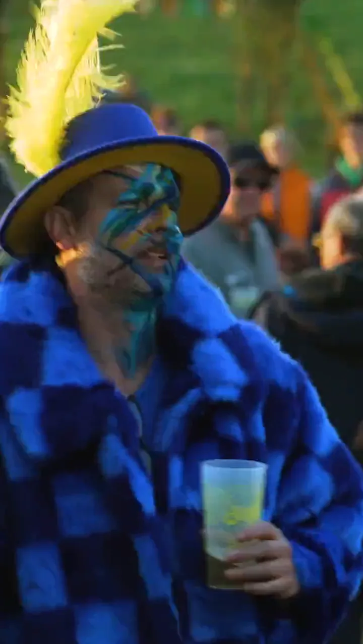 A festival goer with a blue painted face, a feathered hat and a pint in the Solfest crowd