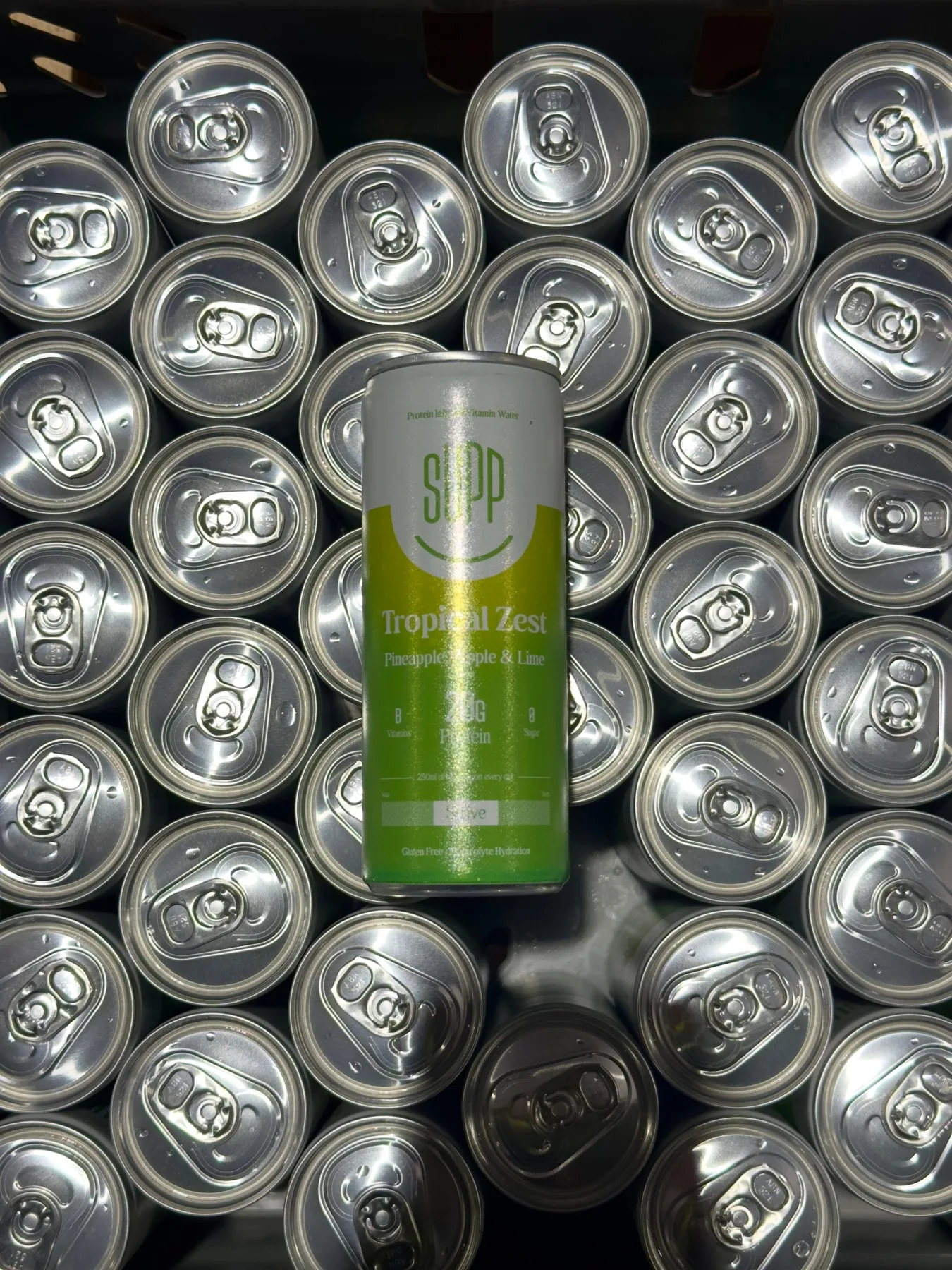 Overhead shot of a Supp Tropical Zest protein water can lying on rows of silver can tops