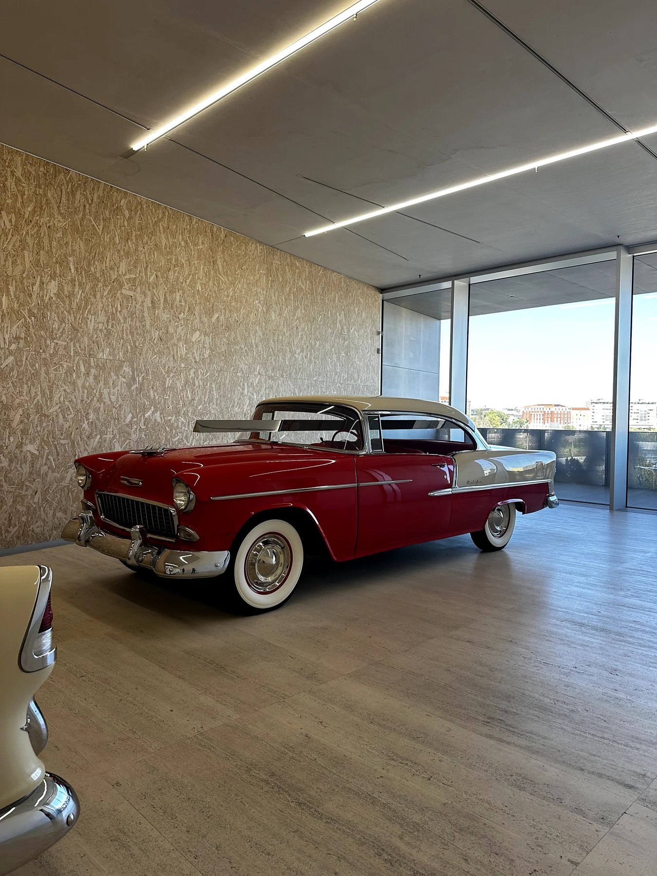 A red and white classic 1950s Chevrolet parked in a bright gallery space with floor to ceiling windows