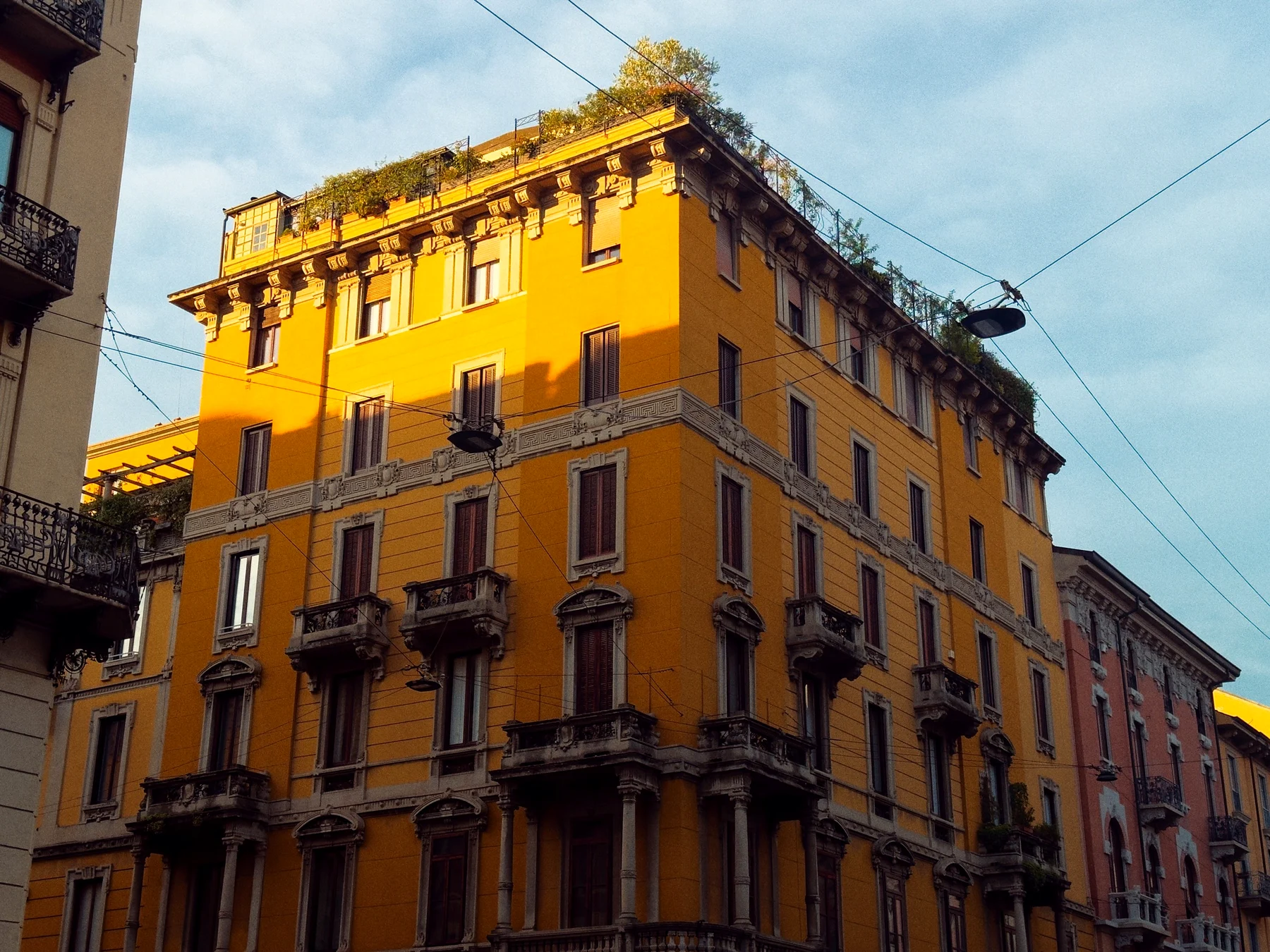 A tall ochre yellow apartment building with green shutters and rooftop plants in Milan, lit by low sun
