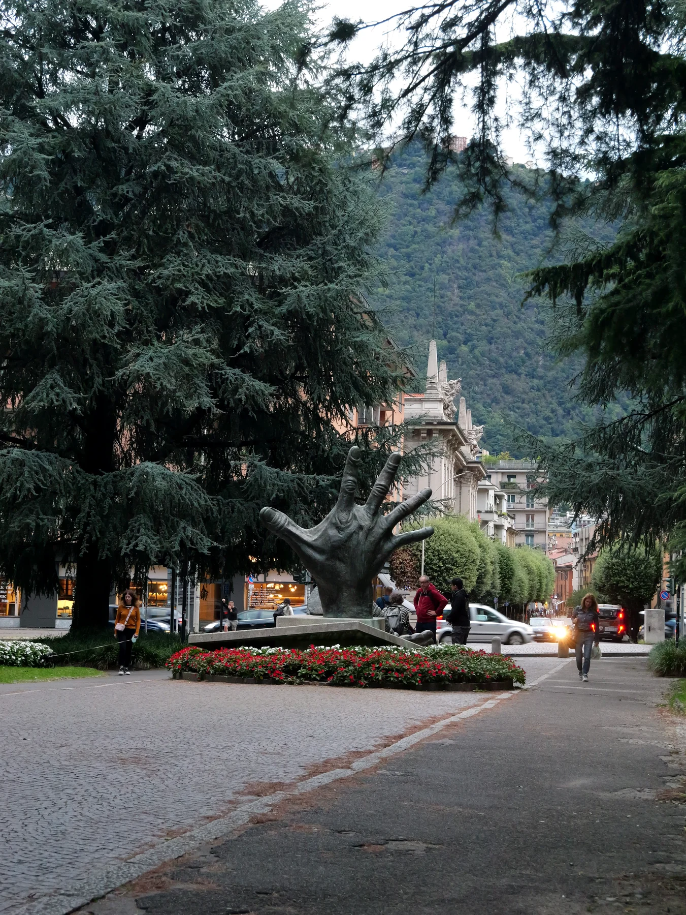 A tree lined promenade at dusk with a giant bronze hand sculpture rising from a bed of red flowers, tall cedars and wooded mountains behind