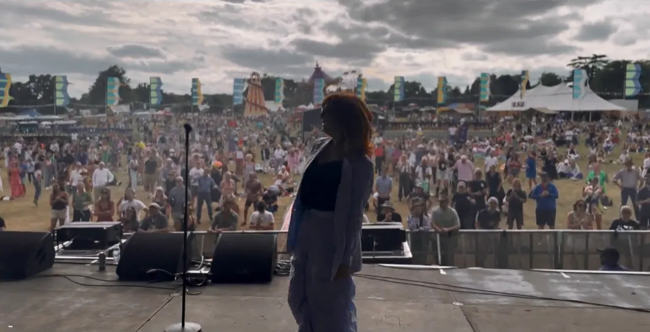 A singer in a grey suit at the front of a festival stage, looking out at a huge crowd under a cloudy sky